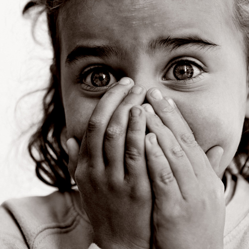 A young girl stares into the camera with her hands over her mouth