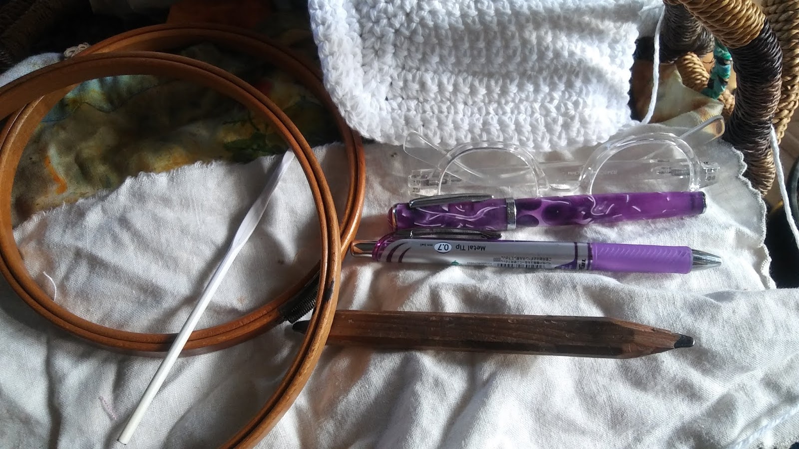 a crochet hook, pencil, and two purple pens rest atop a white cloth. A white, partially crocheted piece sits next to them.