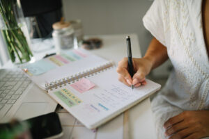 Picture of an open notebook planner on a desk. A woman's arm is holding a pen and writing in it.