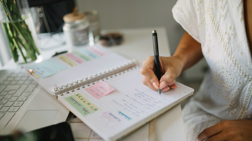 Picture of an open notebook planner on a desk. A woman's arm is holding a pen and writing in it.