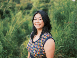 An East Asian woman stands partially facing the camera, smiling. She's in front of a background of greenery and is wearing a dress with a book print.