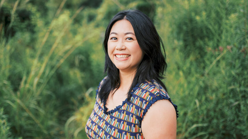 An East Asian woman stands partially facing the camera, smiling. She's in front of a background of greenery and is wearing a dress with a book print.
