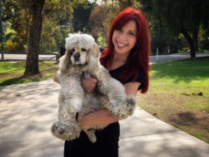 A woman with red hair stands on a path holding a white American Cocker Spaniel