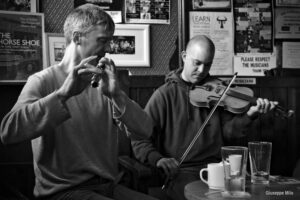 a black and white image of two musicians in an irish pub. Both are wearing sweatshirts. One plays a whistle and one the fiddle.