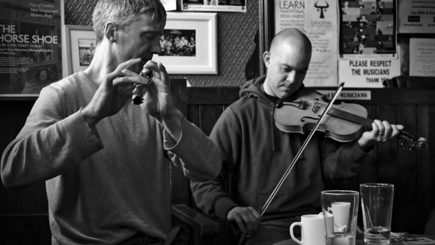 a black and white image of two musicians in an irish pub. Both are wearing sweatshirts. One plays a whistle and one the fiddle.
