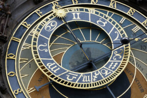 A large astronomical clock takes up the majority of the image. It is primarily a blue-black color with gold outlining. The outer circle of the clock shows the gold numbers of a 24-hour clock, from 8 on the bottom left of the photo to 19 on the top right. There is a smaller circle set atop and in the middle of the larger one which contains the major astrological signs also in gold, while a large clock hand with a golden sun at the end points up towards the top left of the photograph. The title of the photograph is Astronomical Clock by V. Epiney on Flickr.