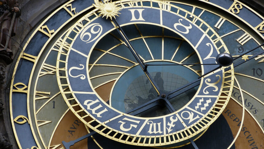 A large astronomical clock takes up the majority of the image. It is primarily a blue-black color with gold outlining. The outer circle of the clock shows the gold numbers of a 24-hour clock, from 8 on the bottom left of the photo to 19 on the top right. There is a smaller circle set atop and in the middle of the larger one which contains the major astrological signs also in gold, while a large clock hand with a golden sun at the end points up towards the top left of the photograph. The title of the photograph is Astronomical Clock by V. Epiney on Flickr.