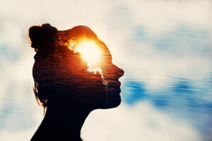 Image of a woman's head in silhouette against a white / blue background, perhaps a sky. Where her brain would be is an image of light, like a sun peeking through the clouds.