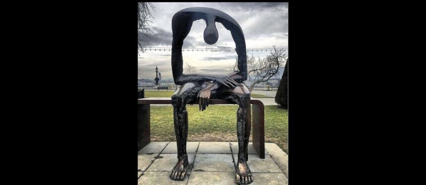 This photo depicts a sculpture of an abstract figure sitting on a bench. The figure is hunched forward with its forearms on its thighs, and there is a massive, empty space in the figure's torso.
