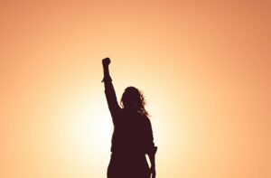 A silhouette of a woman with one arm raised over her head against a warm light background.