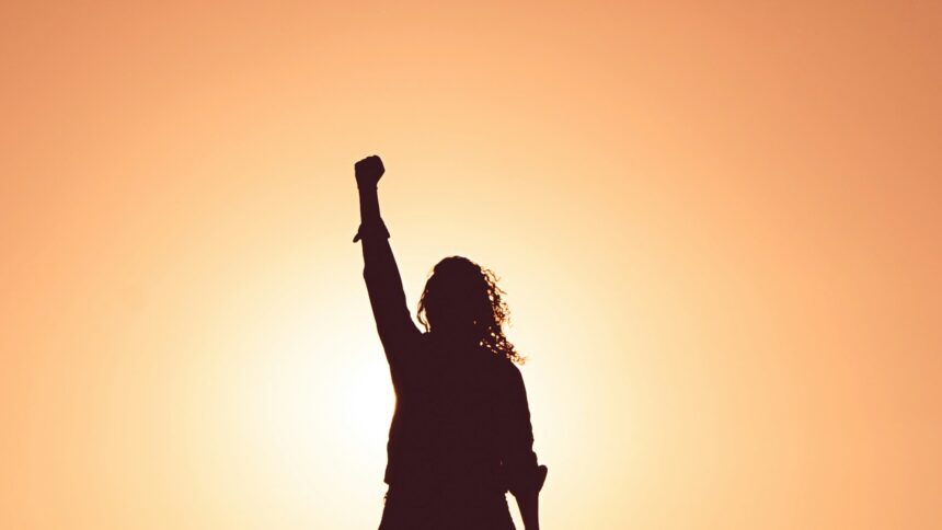 A silhouette of a woman with one arm raised over her head against a warm light background.
