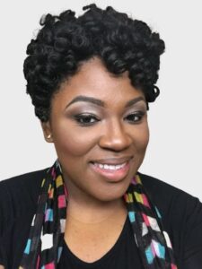 brown-skinned woman with short curly hair, wearing a black sweater and smiling at the camera