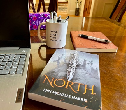 Photo of a laptop on a desk, a mug filled with pens, a notebook, and the novel North by Ann Michelle Harris
